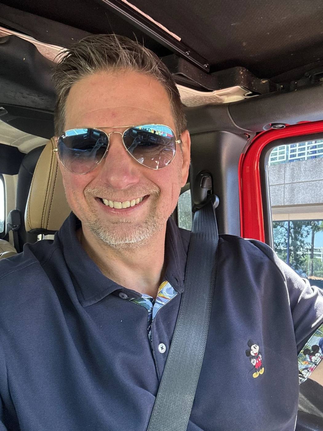 A smiling man with short, dark hair, wearing sunglasses and a dark blue polo shirt with a Mickey Mouse embroidery, sitting in a car with a beige leather seat and a gray seatbelt.
